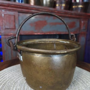 May include: A small, antique brass cauldron with a curved handle. The pot has a rounded body and a slightly flared rim. The metal has a tarnished, aged appearance, suggesting it is vintage. The cauldron sits on a woven mat.