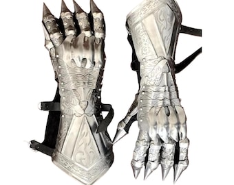 Könnte beinhalten: Ein Paar silberfarbene Metall-Handschuhe mit beweglichen Fingern und scharfen, krallenartigen Spitzen. Die Handschuhe weisen aufwendige eingravierte Designs auf und sind mit schwarzen Lederriemen befestigt. Das Design deutet auf ein mittelalterliches oder Fantasy-Thema hin.