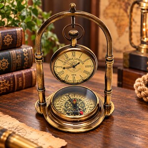 May include: A brass-toned desk clock and compass set featuring an arched frame. The clock face displays Roman numerals, the text Victoria London, and the year 1876. Below the clock, a circular compass features cardinal directions and ornate detailing.