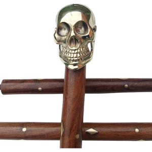 Puede incluir: Bastón decorativo con un mango de calavera de latón. El eje de madera marrón oscuro presenta incrustaciones de latón. El mango de calavera está detallado con cuencas de los ojos, una cavidad nasal y dientes.