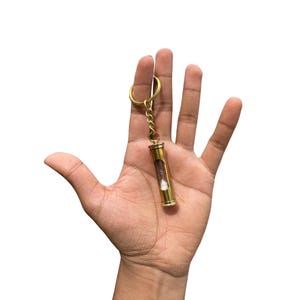 May include: A small, brass hourglass keychain with a gold-colored chain and key ring. The hourglass has a clear glass tube with purple sand. The keychain is held in a hand against a white background.