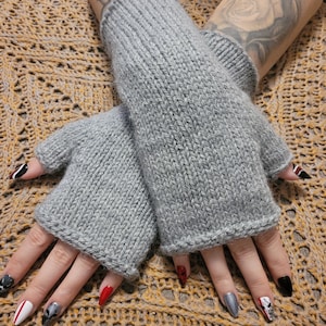 Könnte beinhalten: Ein Paar graue Strick-Fingerhandschuhe. Die Handschuhe sind aus weichem, strukturiertem Garn gefertigt und haben eine gerippte Manschette. Die Handschuhe sind so konzipiert, dass sie die Hände warm halten und gleichzeitig die Finger frei lassen. Ideal für SMS oder andere Aktivitäten.