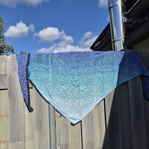 Könnte beinhalten: Ein dreieckiges Häkeltuch mit einem Farbverlauf, der von tiefem Marineblau an den Rändern zu einem helleren Himmelblau und Türkis in der Mitte übergeht. Das Textil weist durchgehend ein komplexes, durchbrochenes Spitzenmuster auf.