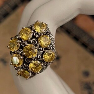 May include: A silver-toned ring featuring a cluster of nine round-cut yellow stones set in a decorative metal frame. The setting includes small, dark, metallic accents nestled between the bright yellow stones, creating a textured, multi-layered appearance.