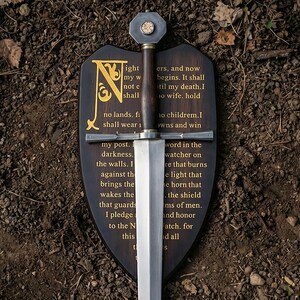 May include: A metal sword with a dark wooden handle and a hexagonal pommel mounted on a dark wooden shield-shaped plaque. The plaque features gold-colored engraved text that begins with a large letter N and includes the Night's Watch oath.