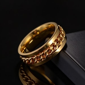 Op de afbeelding: Een goudkleurige ring met een kettingontwerp rond het midden. De ring is gemaakt van glanzend metaal en reflecteert licht. De ring ligt op een zwart oppervlak.