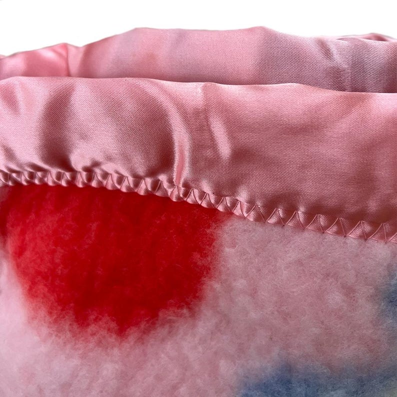 May include: Close-up of a pink and white blanket with a red heart-shaped design. The blanket has a satin border in a matching pink color. The stitching is visible along the edge of the satin.
