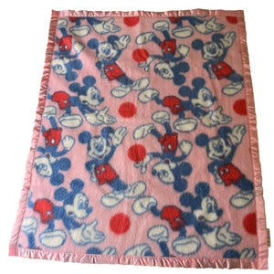 May include: Pink baby blanket featuring a pattern of Mickey Mouse in blue, red, and white. The blanket has a satin edge in a matching pink color. Red polka dots are scattered throughout the design.