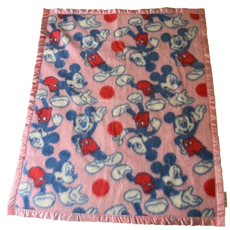 May include: Pink baby blanket featuring a pattern of Mickey Mouse in blue, red, and white. The blanket has a satin edge in a matching pink color. Red polka dots are scattered throughout the design.