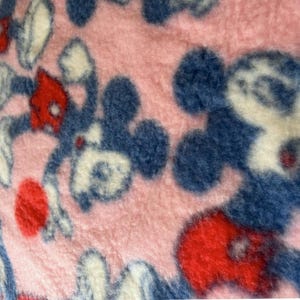 May include: Pink fleece fabric with a repeating pattern of cartoon character figures in blue, red, and white. The fabric has a soft, fuzzy texture, and the cartoon figures are in various poses.