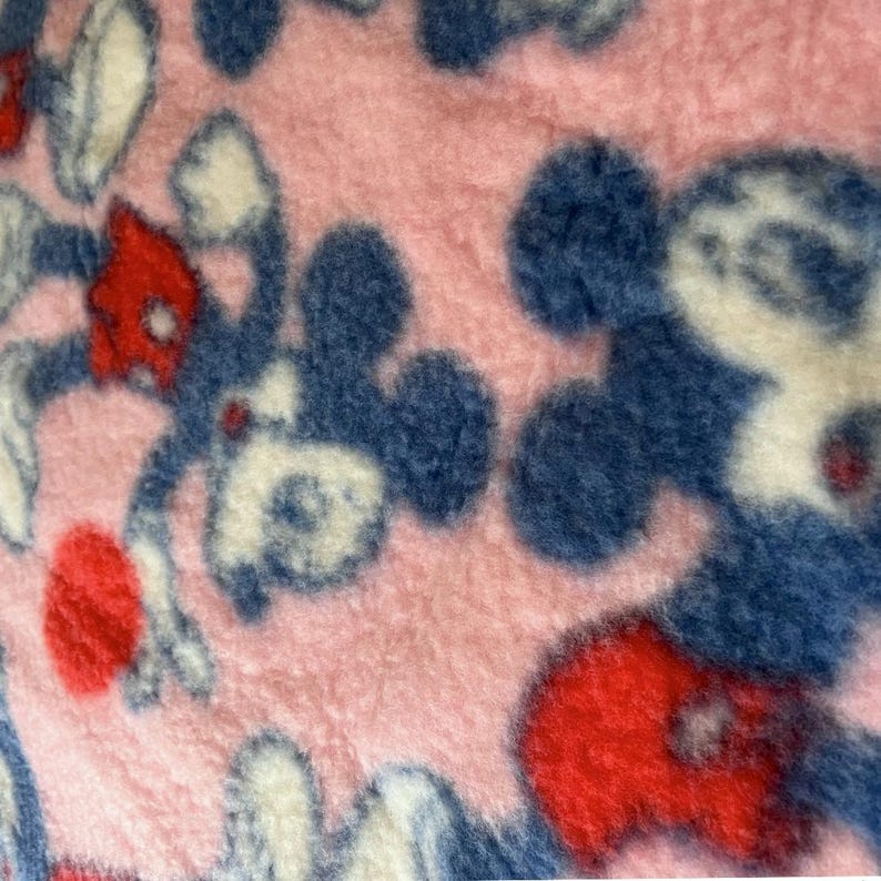 May include: Pink fleece fabric with a repeating pattern of cartoon character figures in blue, red, and white. The fabric has a soft, fuzzy texture, and the cartoon figures are in various poses.
