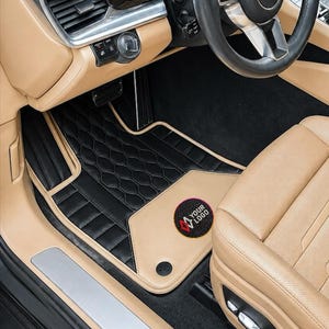 May include: A black car floor mat with a hexagonal quilted pattern and a tan border, featuring a circular patch with the text YOUR LOGO. The mat is installed in a vehicle interior with tan leather seats, a black dashboard, and a black steering wheel.