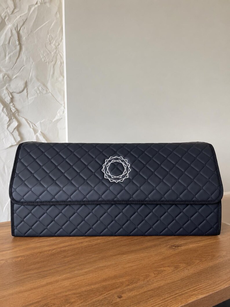 Op de afbeelding: Een marineblauwe, gewatteerde opbergorganizer met een geometrisch patroon en een decoratief wit embleem. De organizer is rechthoekig met een scharnierend deksel en staat op een houten oppervlak.