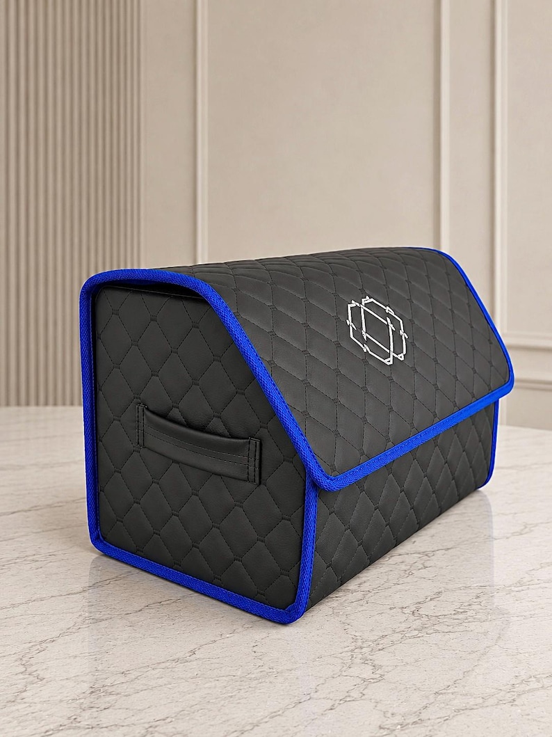 Op de afbeelding: Zwarte gewatteerde opbergcontainer met blauwe rand en een geometrisch ontwerp op het deksel. De container heeft een handvat en staat op een marmeren oppervlak. Ontworpen voor opslag.