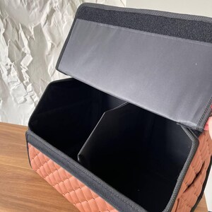 Op de afbeelding: Een bruine, gewatteerde auto-organizer met zwarte rand en een verdeeld interieur. De organizer heeft een rechthoekige vorm en een deksel dat opent om opbergvakken te onthullen. De binnenkant is zwart en de buitenkant is warmbruin.