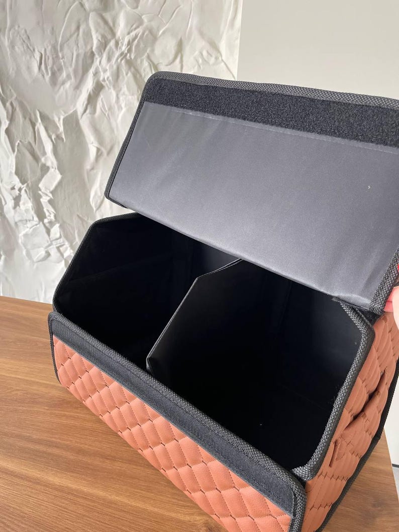 Op de afbeelding: Een bruine, gewatteerde auto-organizer met zwarte rand en een verdeeld interieur. De organizer heeft een rechthoekige vorm en een deksel dat opent om opbergvakken te onthullen. De binnenkant is zwart en de buitenkant is warmbruin.