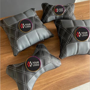 May include: A collection of black and gray car seat headrest pillows with white stitching in a diamond pattern. Each pillow features a circular logo with the text "YOUR LOGO" in a colorful design.