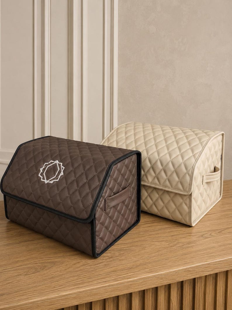 Op de afbeelding: Twee gewatteerde opbergdozen. E&eacute;n is bruin met een wit geometrisch ontwerp en zwarte rand. De andere is beige. Beide hebben handvatten en diamantstiksels. Ze staan op een houten oppervlak.