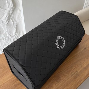 Op de afbeelding: Zwarte rechthoekige opbergdoos met een gewatteerd patroon en een geometrisch ontwerp in het midden. De doos heeft een handvat aan de zijkant en staat op een houten oppervlak.