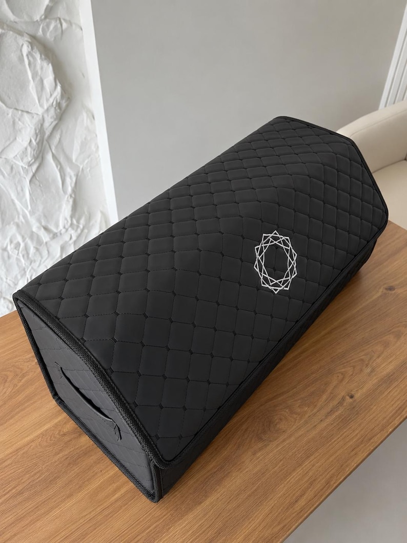 Op de afbeelding: Zwarte rechthoekige opbergdoos met een gewatteerd patroon en een geometrisch ontwerp in het midden. De doos heeft een handvat aan de zijkant en staat op een houten oppervlak.