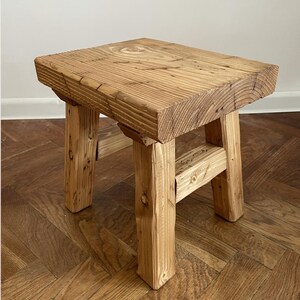 Peut inclure: Un petit tabouret en bois rustique avec une assise rectangulaire et quatre pieds robustes. Le grain naturel du bois est visible partout, mettant en valeur les nœuds et les textures du matériau. Le tabouret mesure environ 30 cm de haut et convient à divers usages.