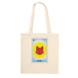 Puede incluir: Una bolsa de tela beige con un estampado gráfico rectangular de una cara de gato roja centrada dentro de un sol amarillo brillante. El diseño incluye el número 46 en la esquina superior izquierda y el texto EL SOL en la parte inferior.