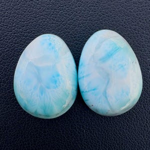 Può includere: Due pietre preziose di Larimar levigate a forma di goccia con una superficie liscia e lucida. Le pietre presentano un motivo marmorizzato nei toni dell'azzurro cielo, turchese e bianco, con venature complesse simili a nuvole sulle forme opache.
