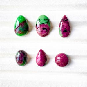 May include: Six polished cabochon gemstones arranged in two rows. The stones feature a mix of vibrant magenta, deep red, and bright green colors with black inclusions. The shapes include ovals, teardrops, and a circle, all with a smooth finish.