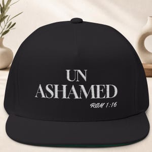 May include: A black baseball cap featuring white embroidered text that reads UN ASHAMED with the smaller text ROM 1:16 positioned underneath. The cap has a structured crown, a flat brim, and a button at the top centre of the crown.