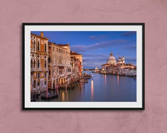 May include: Framed print of a scenic view of Venice, Italy, featuring buildings along a canal and a domed church in the distance. The water reflects the buildings and the sky, creating a serene atmosphere. The sky is a gradient of blue and orange.