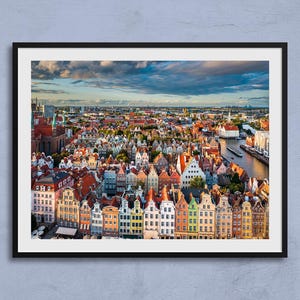 Puede incluir: Una impresión enmarcada que muestra un vibrante paisaje urbano. La imagen captura una densa colección de edificios coloridos con tejados rojos, naranjas y blancos. Un río serpentea por la ciudad y el cielo está lleno de nubes dramáticas.