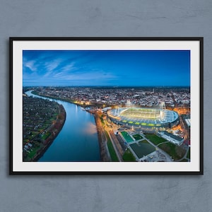 Puede incluir: Fotografía aérea de un estadio iluminado, situado cerca de un río y una ciudad al anochecer. La imagen está enmarcada en un borde negro, con un cielo azul y nubes en el fondo.