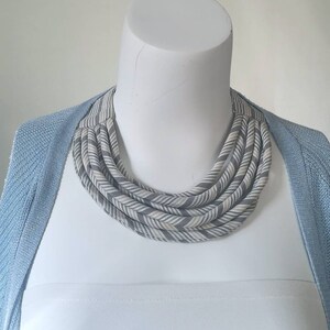 May include: A gray and white striped fabric necklace with a chevron pattern. The necklace is layered and has a chunky, bold look.