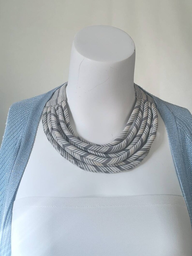 May include: A gray and white striped fabric necklace with a chevron pattern. The necklace is layered and has a chunky, bold look.