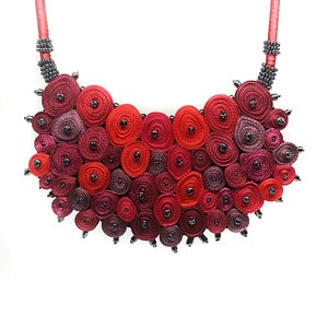 May include: A red and burgundy statement necklace made of rolled fabric and black beads. The necklace is shaped like a crescent moon and is a bold and eye-catching piece of jewelry.