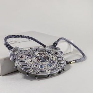 May include: A handcrafted necklace with a large, oval pendant. The pendant features intricate beadwork in shades of gray, lavender, and cream, with accents of blue and black. The necklace has a gray cord and a gold-colored clasp.