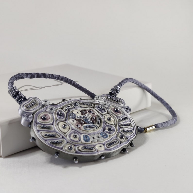 May include: A handcrafted necklace with a large, oval pendant. The pendant features intricate beadwork in shades of gray, lavender, and cream, with accents of blue and black. The necklace has a gray cord and a gold-colored clasp.