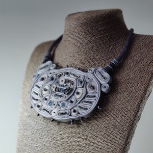 May include: A handcrafted statement necklace in shades of blue, white, and gray. The intricate design features a large, oval-shaped pendant with detailed patterns and bead accents. The necklace is displayed on a mannequin.