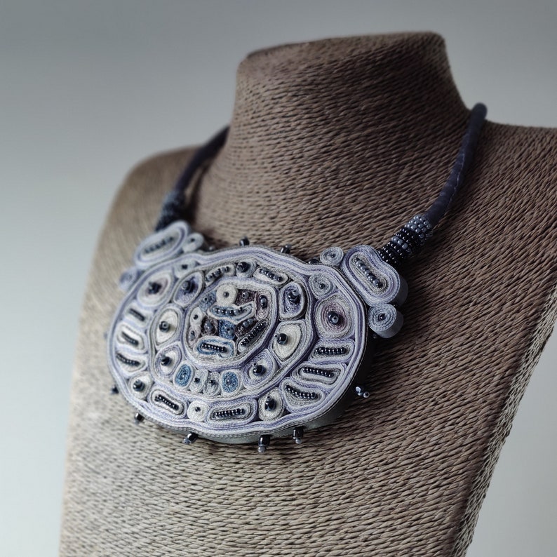 May include: A handcrafted statement necklace in shades of blue, white, and gray. The intricate design features a large, oval-shaped pendant with detailed patterns and bead accents. The necklace is displayed on a mannequin.