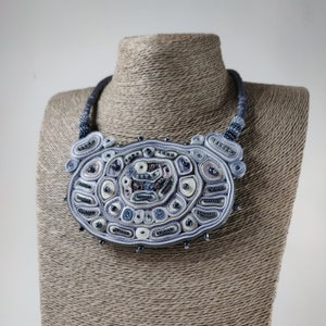 May include: A handcrafted statement necklace with a large, oval pendant. The pendant features intricate beadwork in shades of blue, white, and gray, with a textured, layered design. The necklace is displayed on a mannequin.