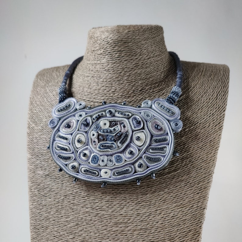 May include: A handcrafted statement necklace with a large, oval pendant. The pendant features intricate beadwork in shades of blue, white, and gray, with a textured, layered design. The necklace is displayed on a mannequin.