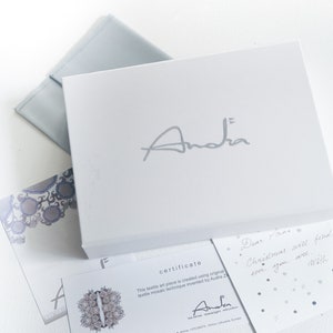 May include: A white box with the name "Andra" in silver lettering, accompanied by a light blue envelope and several cards. One card reads "certificate" and another has a handwritten note. The overall aesthetic is clean and elegant.