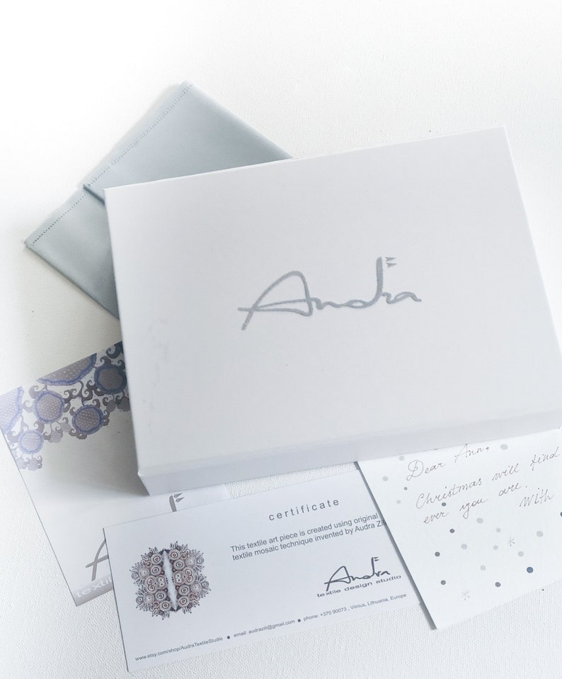May include: A white box with the name "Andra" in silver lettering, accompanied by a light blue envelope and several cards. One card reads "certificate" and another has a handwritten note. The overall aesthetic is clean and elegant.