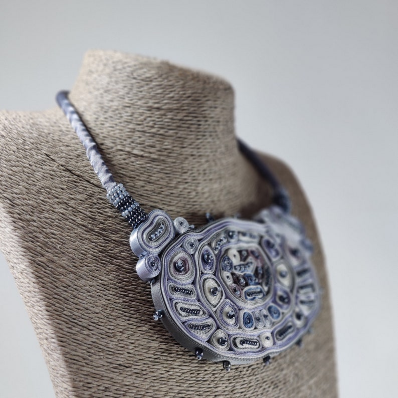 May include: A handcrafted statement necklace in shades of lavender, gray, and white. The intricate design features a central circular pendant with layered textures and bead accents, displayed on a mannequin.