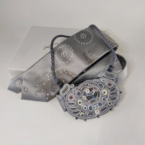May include: A handcrafted necklace with a large, oval pendant in shades of gray, blue, and white. The pendant is intricately detailed with various shapes and textures. The necklace rests on a gray tie with a subtle pattern, next to a white box.