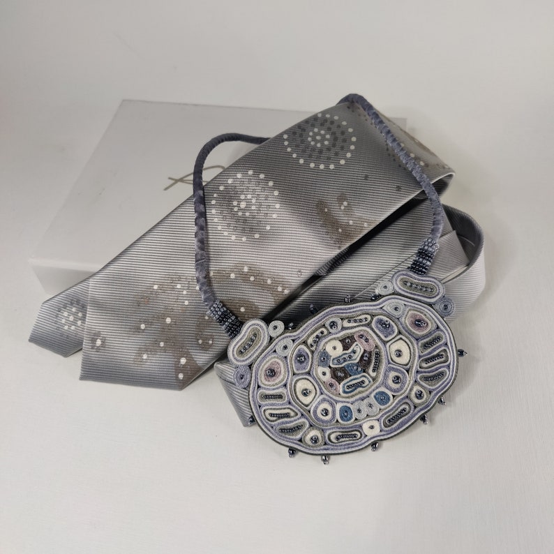 May include: A handcrafted necklace with a large, oval pendant in shades of gray, blue, and white. The pendant is intricately detailed with various shapes and textures. The necklace rests on a gray tie with a subtle pattern, next to a white box.