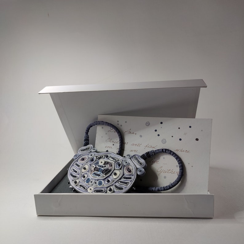 May include: A handcrafted, oval-shaped purse with a beaded design in shades of blue, white, and black, presented in a white gift box. The purse has two circular handles. A handwritten card is inside the box.