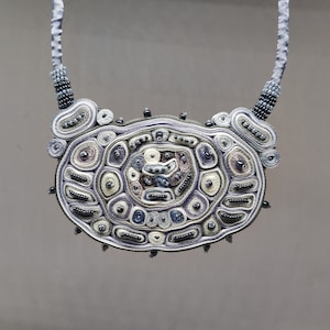 May include: A handcrafted necklace featuring a large, oval pendant with intricate, layered designs in shades of white, gray, and purple. The pendant is accented with small, dark beads and attached to a light gray ribbon.