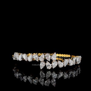 May include: A gold bracelet with a row of pear-shaped diamonds. The diamonds are set closely together, creating a continuous sparkle. The bracelet is displayed on a black surface, reflecting the diamonds' brilliance. The overall design is elegant and luxurious.