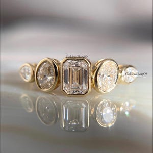 May include: A gold ring featuring a series of bezel-set diamonds in various shapes: pear, oval, emerald cut, and round. The diamonds are clear and sparkling, set against a polished gold band. The ring is displayed on a reflective surface.